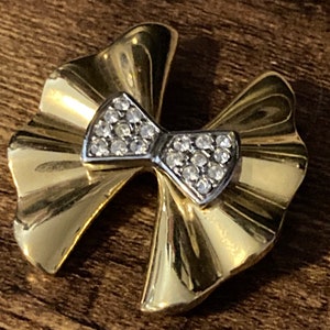 May include: A gold-toned brooch in the shape of a bow with clear rhinestones. The bow is set on a gold-toned, leaf-shaped base.
