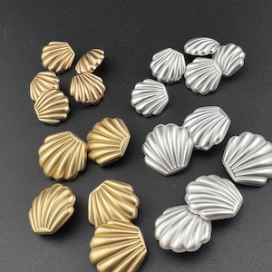 May include: A selection of seashell-shaped decorative elements in gold and silver hues. The elements are arranged on a dark surface, highlighting their detailed, ridged texture. These embellishments are suitable for various craft projects.