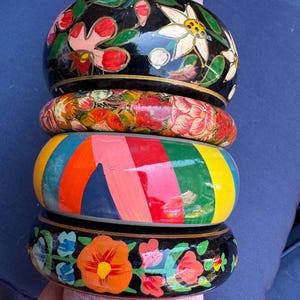 May include: Four wooden bangles stacked together. The top bangle is black with white and pink floral patterns. The second bangle is red with floral designs. The third bangle is rainbow-striped. The bottom bangle is black with orange and blue floral designs.