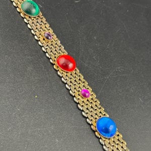 Könnte beinhalten: Goldfarbenes Armband mit einem gewebten Kettendesign. Das Armband hat fünf farbige, oval geformte Edelsteine: grün, lila, rot, lila und blau. Der Verschluss ist ein einfacher Haken- und Ösenverschluss.