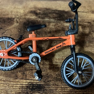 Op de afbeelding: Oranje miniatuur mountainbike met zwarte banden en een zwart zadel. De fiets heeft de tekst "Mountain Bike" op het frame. De wielen zijn zilverkleurig met zwarte banden. Een speelgoed.