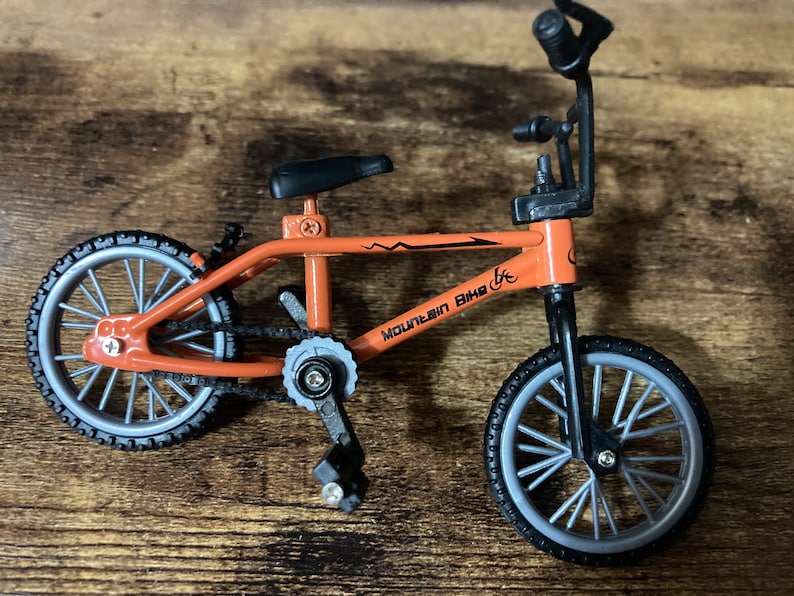 Op de afbeelding: Oranje miniatuur mountainbike met zwarte banden en een zwart zadel. De fiets heeft de tekst "Mountain Bike" op het frame. De wielen zijn zilverkleurig met zwarte banden. Een speelgoed.