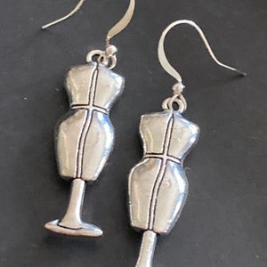 Könnte beinhalten: Ein Paar silberfarbene Ohrringe in Form von Miniatur-Schneiderpuppen. Die Ohrringe haben einen Hakenverschluss und ein detailliertes Vintage-Design. Die Schneiderpuppen haben ein Kreuzstichmuster auf der Vorderseite.