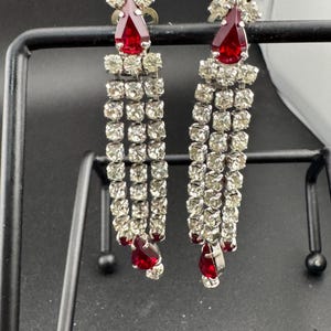 May include: A pair of vintage chandelier earrings. Each earring has a teardrop-shaped, ruby-red gemstone at the top and bottom, with multiple rows of clear, sparkling rhinestones cascading down. The earrings are displayed on a black metal stand.