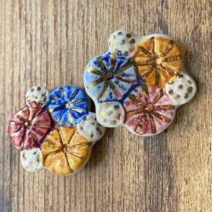 May include: Two ceramic buttons featuring a floral design. The buttons are white with gold, blue, and red accents. The buttons are shaped like flowers with multiple petals.