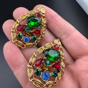 May include: A pair of vintage clip-on earrings. Each earring features a gold-toned, teardrop-shaped frame with an intricate design. The earrings are adorned with a large green oval stone, red teardrop-shaped stones, and smaller green and blue round stones.