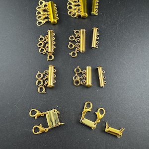 May include: Various gold-coloured jewellery clasps and connectors, including multi-strand clasps with lobster claw closures and cylindrical connectors with multiple loops. The clasps are arranged on a dark surface, highlighting their shiny finish.