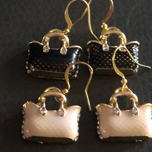 Puede incluir: Un par de pendientes de oro en forma de bolso de mano. Un par es negro con detalles dorados y el otro par es blanco con detalles dorados.