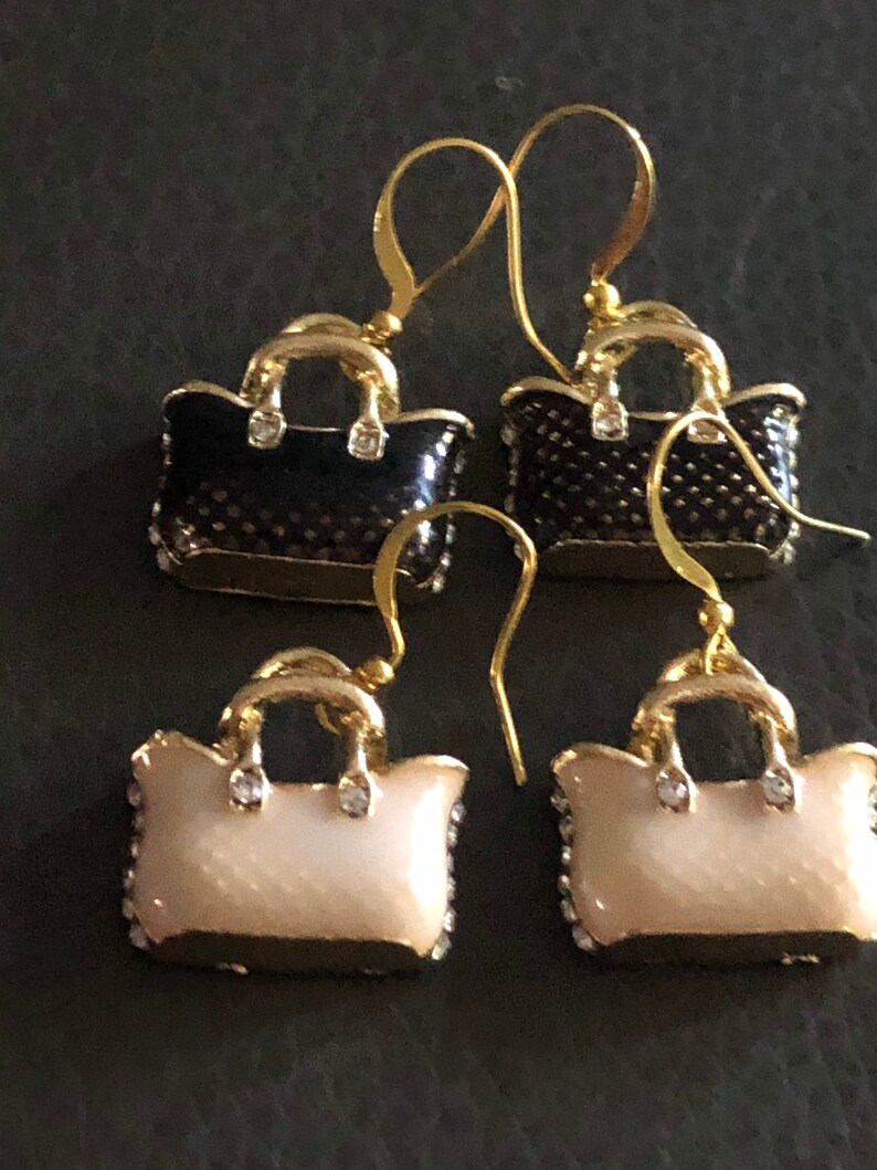 Puede incluir: Un par de pendientes de oro en forma de bolso de mano. Un par es negro con detalles dorados y el otro par es blanco con detalles dorados.