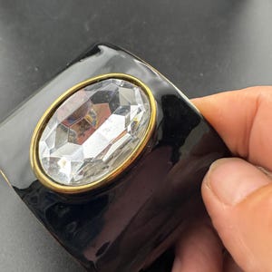 以下が含まれることがあります： 光沢のある黒い仕上げの幅広のバングルブレスレット。中央にはゴールドトーンの金属ベゼルにセットされた、大きな楕円形の透明なファセットカットクリスタルがあしらわれています。ブレスレットは滑らかで反射性のある表面を持ち、大胆で構造的なデザインが特徴です。