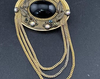 May include: Vintage oval brooch with a black center stone, surrounded by gold-tone metal and dangling chains. The brooch features leaf accents and small, iridescent rhinestones. A classic piece of jewelry.