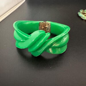 May include: A bright green hinged bangle bracelet featuring a thick, twisted, sculptural design. The bracelet is constructed from a smooth, glossy material and includes a metallic gold-tone hinge mechanism that allows the piece to open and close.