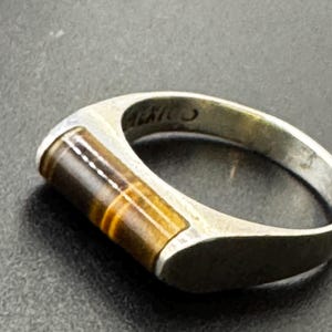 以下が含まれることがあります： 茶色と金色のタイガーアイの宝石が長方形にはめ込まれた銀色の金属製リング。バンドは滑らかで光沢のある仕上げで、金属バンドの内側には「MEXICO」という文字がはっきりと刻印されています。
