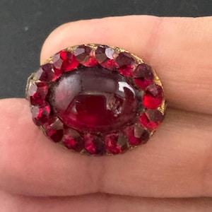 May include: An antique oval-shaped brooch with a central, deep red gemstone surrounded by smaller, faceted red stones. The setting is a gold-toned metal, and the brooch is held in a hand.