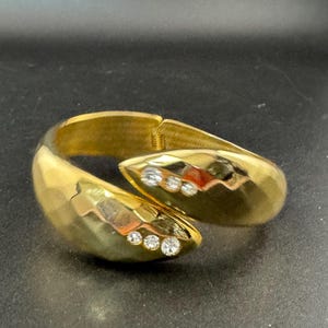 May include: A gold-coloured hinged cuff bracelet with a textured surface. The bracelet features three clear, round gemstones on each end. The bracelet is designed to wrap around the wrist.