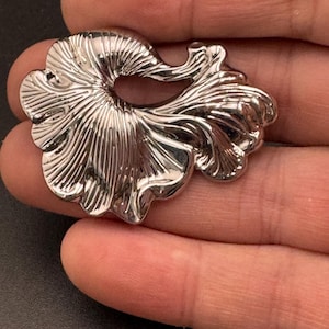 May include: A shiny, silver-coloured metal brooch with a floral design. The brooch features a detailed, textured pattern with a central opening and flowing, petal-like shapes. The design is highly reflective, catching the light.