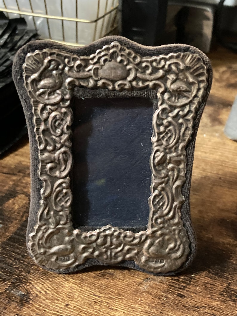 Puede incluir: Un marco de fotos plateado con un dise&ntilde;o ornamentado y un respaldo de terciopelo azul oscuro.