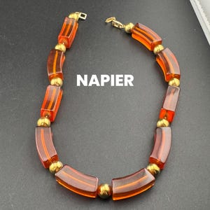 May include: A vintage Napier necklace with translucent orange rectangular beads and gold accents. The necklace has a gold-tone clasp. The word "NAPIER" is printed on the image. The necklace is displayed on a dark surface.