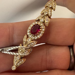 May include: A gold-toned brooch featuring a central oval ruby surrounded by clear crystals. The brooch has a leaf-like design with additional crystals, and is attached to a white string.