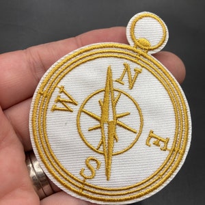 May include: Gold embroidered compass patch with a white background and a gold loop at the top. The compass points north, south, east, and west.