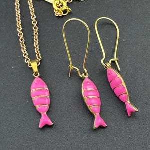 May include: A gold-coloured necklace and earring set featuring pink fish pendants. The fish pendants have gold-coloured accents and are suspended from gold-coloured chains and earring hooks. The jewellery set is displayed on a dark background.