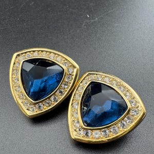 May include: A pair of gold-toned earrings with a triangular design. Each earring features a large, deep blue gemstone at the center, surrounded by a border of small, clear crystals. The earrings have a classic, elegant style.