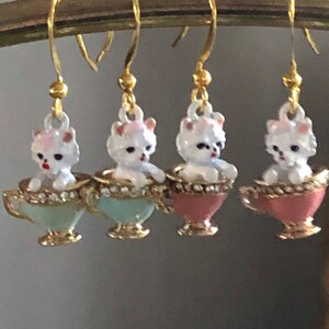 Puede incluir: Cuatro pares de pendientes colgantes con un perro de cer&aacute;mica blanco sentado en una taza de t&eacute;. Las tazas de t&eacute; son de tres colores: azul claro, rosa y rosa oscuro.