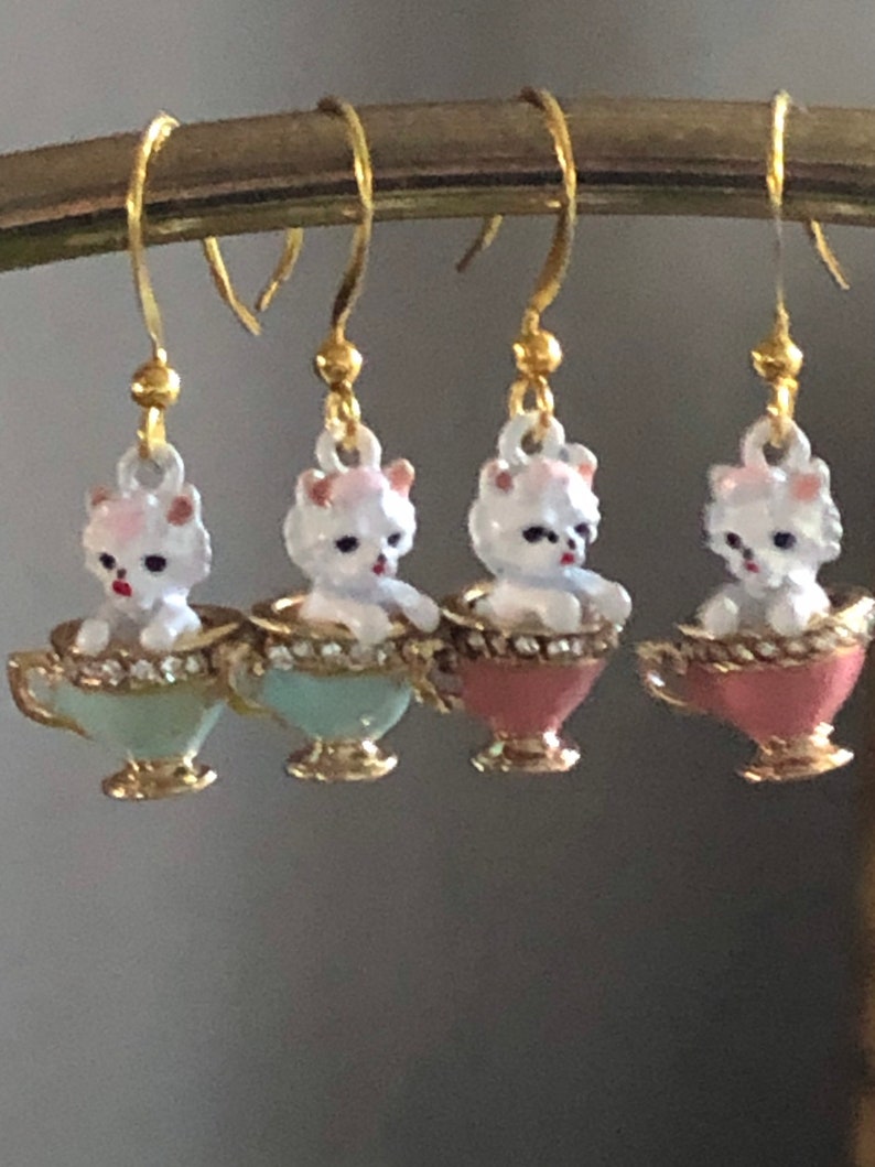 Puede incluir: Cuatro pares de pendientes colgantes con un perro de cer&aacute;mica blanco sentado en una taza de t&eacute;. Las tazas de t&eacute; son de tres colores: azul claro, rosa y rosa oscuro.