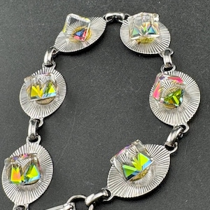 May include: A silver bracelet with iridescent, square crystals set on oval, sunburst-patterned links. The bracelet has a clasp and is designed for wrist wear. The crystals reflect a range of colours.