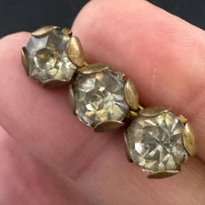 Peut inclure: Gros plan sur une broche vintage dorée. Elle présente trois pierres de cristal claires et facettées, chacune sertie dans un décor en métal doré. La broche est tenue dans la main d'une personne sur fond sombre.