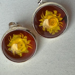 May include: A pair of round stud earrings. Each earring features a yellow rose encased in a clear, domed resin set against a dark red background. The earrings have a silver-colored metal border and post.