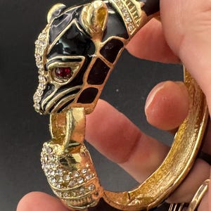 May include: A gold-tone hinged bangle bracelet featuring a stylized panther head design. The panther is decorated with black enamel, clear rhinestones along the snout and collar, and a single red rhinestone eye. The metal has a textured finish.
