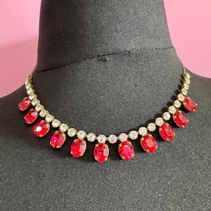 May include: A gold-coloured necklace featuring alternating red oval gemstones and clear round rhinestones. The necklace is displayed on a black mannequin head against a pink background. The red stones are evenly spaced.