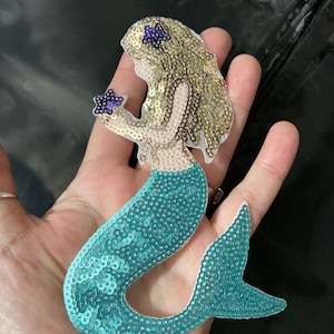 May include: A turquoise and gold sequin mermaid applique with a purple star in her hand.