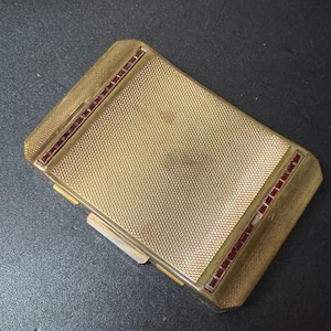 May include: A gold-colored compact with a textured surface and a border of red rhinestones.