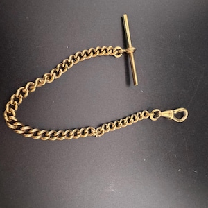 May include: A gold-coloured metal watch chain featuring a curb link design. The chain is equipped with a T-bar at one end and a spring-loaded swivel clasp at the opposite end, providing a secure attachment point for a pocket watch or accessory.