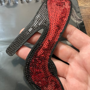 Peut inclure: Patch thermocollant en forme de chaussure à talon haut rouge et noir. La chaussure est faite de paillettes et a un contour noir.