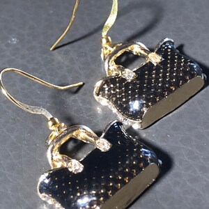 Puede incluir: Un par de pendientes colgantes de tono dorado con un dise&ntilde;o de bolso negro. El bolso tiene un asa de tono dorado y diamantes de imitaci&oacute;n.