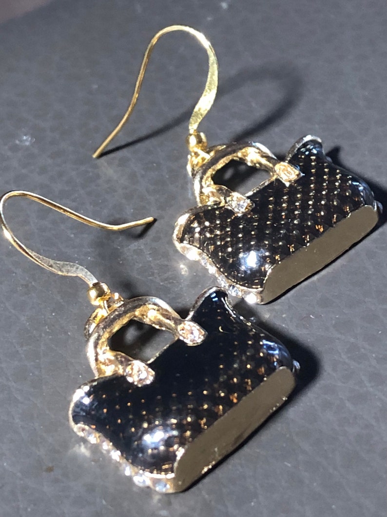 Puede incluir: Un par de pendientes colgantes de tono dorado con un dise&ntilde;o de bolso negro. El bolso tiene un asa de tono dorado y diamantes de imitaci&oacute;n.