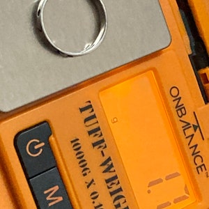 Może przedstawiać: Zbliżenie na cyfrową wagę z pomarańczową obudową. Waga wskazuje "1,1" grama. Waga jest oznaczona jako "Tuff-Weigh" i "1000g x 0,1g".