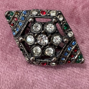 May include: A vintage brooch with a diamond shape, featuring a central cluster of clear rhinestones surrounded by smaller clear, red, blue, and green stones. The metal has an antique silver finish, set against a pink background.