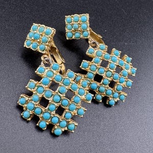 May include: A pair of gold-coloured earrings with a square top and a geometric, openwork design. Each earring features small, round, turquoise-coloured stones set within the gold frame. The earrings are displayed against a dark background.