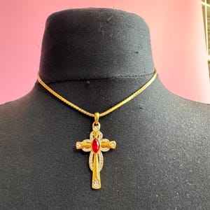 Peut inclure: Collier avec pendentif en forme de croix dorée, orné d'une pierre rouge au centre et de pierres claires. La croix est suspendue à une fine chaîne dorée. Le collier est présenté sur un mannequin noir.
