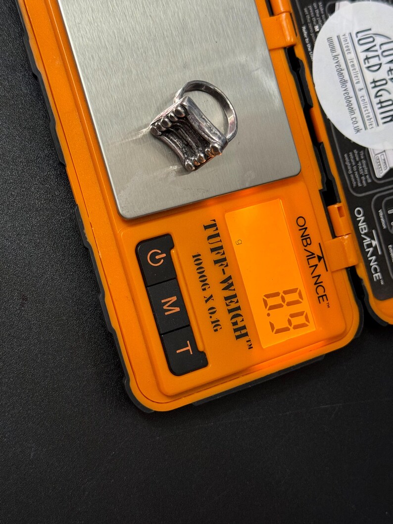 May include: A silver ring with a textured design, resembling a stack of books, is displayed on a digital scale. The scale reads 8.9 grams.