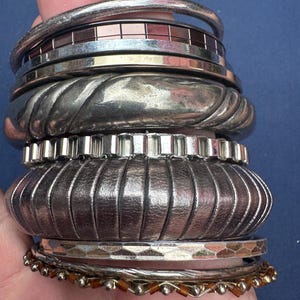 May include: A stack of silver-toned metal bracelets featuring various textures and designs, including smooth bands, a wide ribbed cuff, a segmented metallic piece, a hammered finish band, and a thin bracelet with small amber-coloured beads.