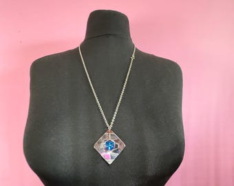 May include: A silver necklace with a geometric pendant. The pendant is a diamond shape with a blue center and pink and silver accents. The necklace is displayed on a black mannequin against a pink background.