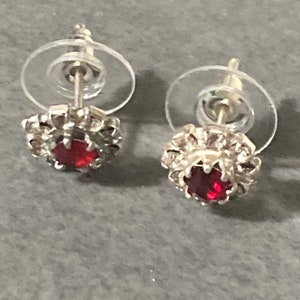 Puede incluir: Un par de pendientes de bot&oacute;n de plata con un centro de piedra roja y un respaldo de pl&aacute;stico transparente.