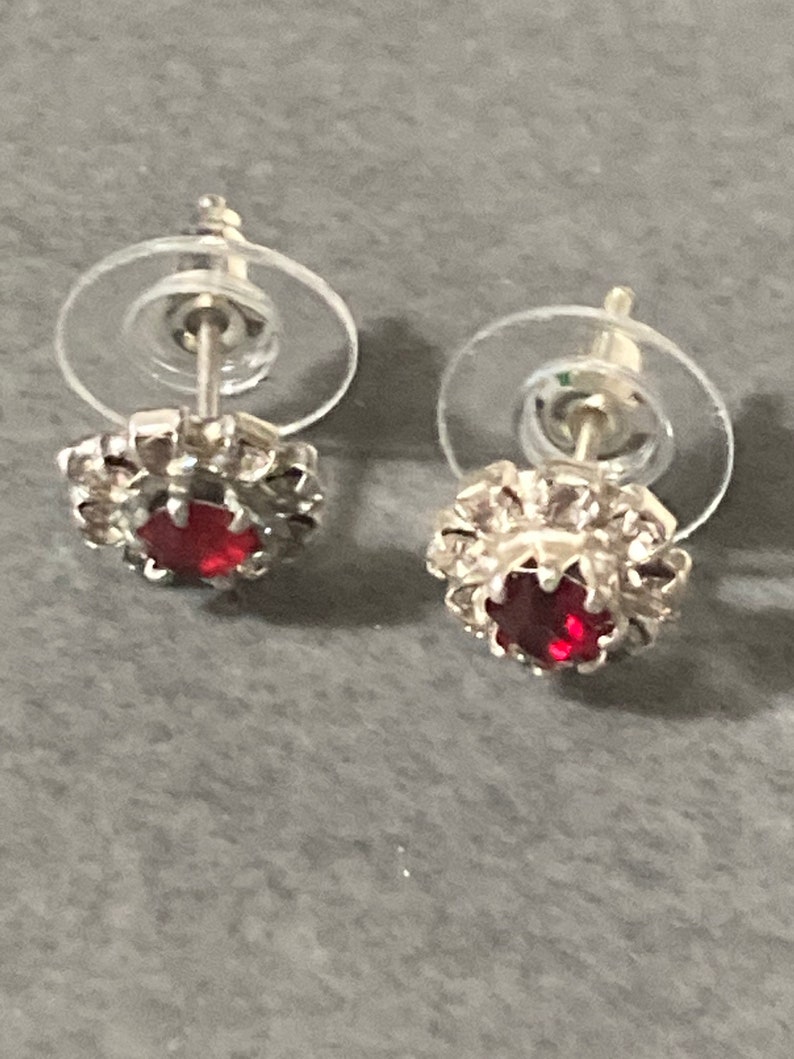 Puede incluir: Un par de pendientes de bot&oacute;n de plata con un centro de piedra roja y un respaldo de pl&aacute;stico transparente.