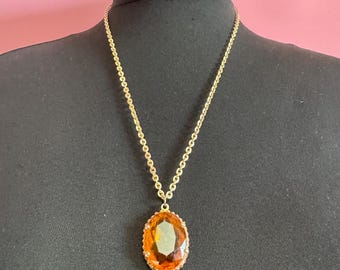 May include: A gold-toned necklace with a large, oval amber-colored pendant. The pendant is set in a gold frame and hangs from a delicate chain. The necklace is displayed on a black mannequin.
