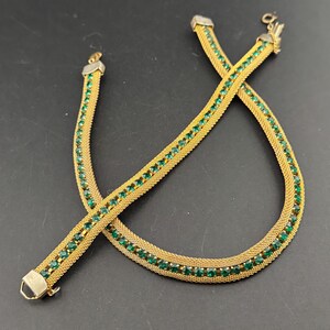 May include: A gold-toned necklace with a mesh design, adorned with rows of small, emerald-green gemstones. The necklace is laid out on a dark surface, showcasing its intricate details and clasp. The jewelry piece appears to be vintage or antique.